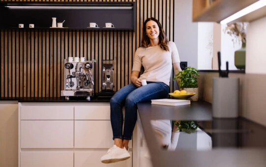 Glückliche junge Frau sitzt auf Danküche mit hellen Fronten und dunkler Arbeitsplatte und hält eine Espresso Tasse in der Hand. 