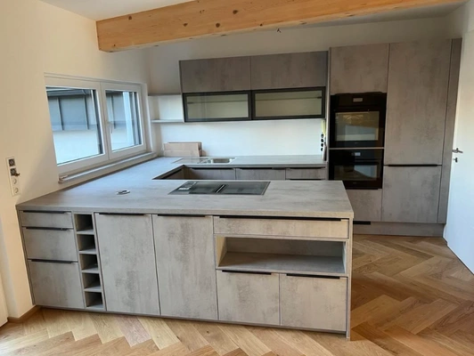 Kundenküche in Oberndorf Kundenküche im Modell Andros, mit Fronten in Beton, Griffleisten in Schwarz und zwei Hängeschränken mit Glasfronten.