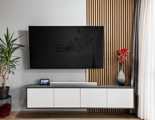 Weißes TV Lowboard mit Ferseher darauf, dahinter moderne Holzlamellen als dekoratives Element.