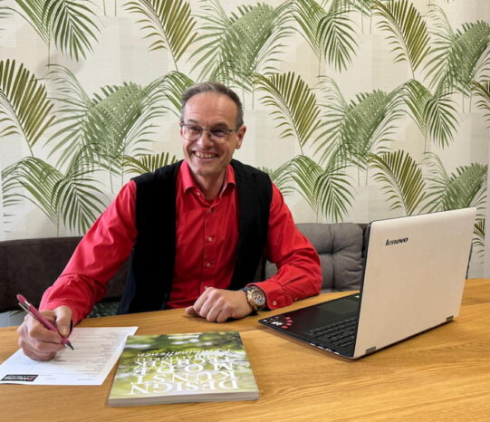 Dietmar Haring sitzt am Küchenplanungstisch mit einem Katalog und einen Laptop. 