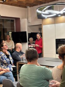Dietmar Haring begrüßt die Gäste zu einer Kochshow im DANKÜCHEN Küchenstudio. Mehrere Besucher sitzen vor einer modernen Ausstellungsküche und hören der Begrüßung zu.